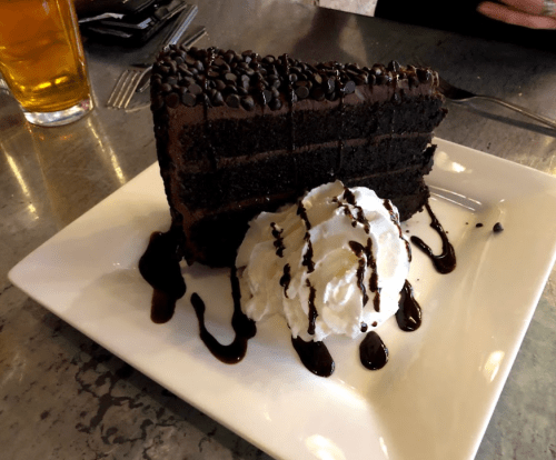 A slice of rich chocolate cake topped with chocolate chips, served with whipped cream and chocolate drizzle on a plate.