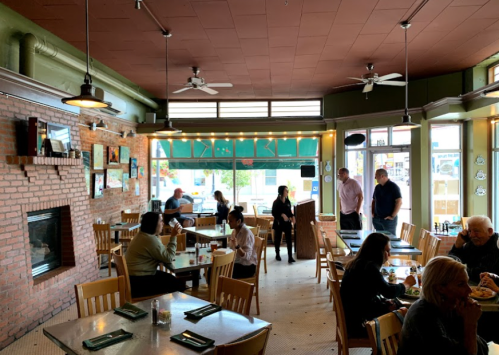 Cozy restaurant interior with brick walls, wooden tables, and patrons dining. Large windows let in natural light.