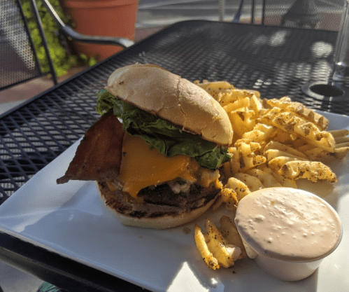 A juicy burger with bacon, cheese, and lettuce, served with crinkle-cut fries and dipping sauce on a white plate.