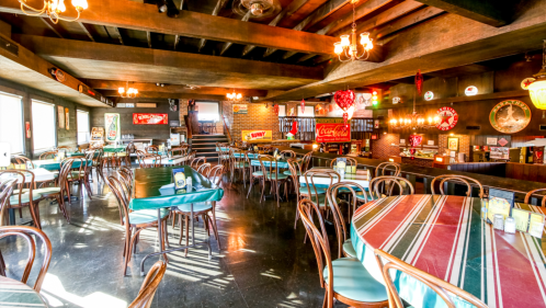 Cozy restaurant interior with wooden beams, tables set with green and red tablecloths, and a bar in the background.