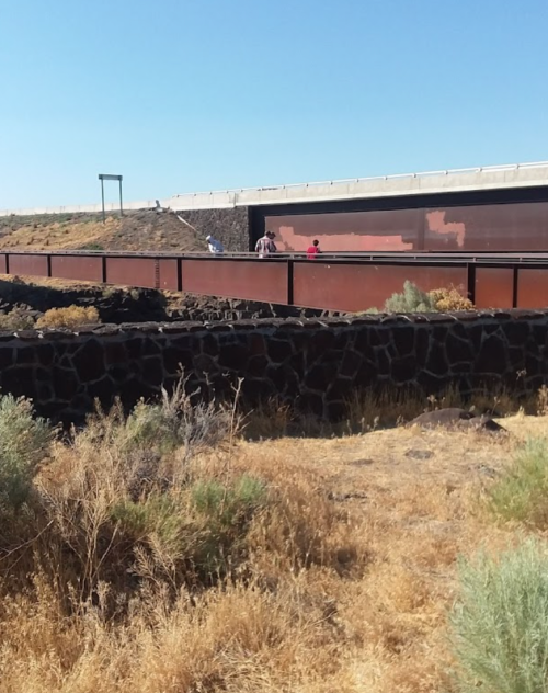 A view of a border wall with people walking nearby, surrounded by dry grass and rocky terrain under a clear blue sky.