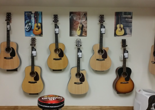 A wall display of various acoustic guitars with price tags, alongside posters of guitars. A round container is in the foreground.