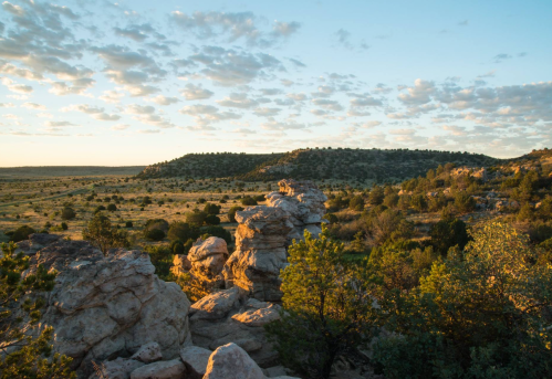 A scenic landscape featuring rocky formations, green vegetation, and a vast sky with scattered clouds at sunset.