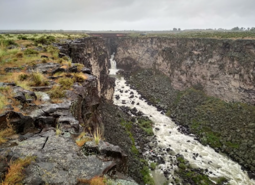 A dramatic canyon with steep rocky cliffs and a waterfall cascading into a turbulent river below, under a cloudy sky.