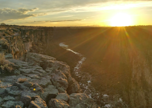 Sunset over a canyon with a winding river, casting warm light on rocky cliffs and the surrounding landscape.