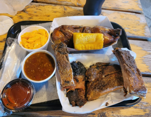 A tray of barbecue featuring ribs, chicken, macaroni and cheese, cornbread, and a cup of sauce.