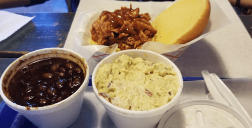 A plate featuring pulled pork with barbecue sauce, a bun, and two sides: black beans and coleslaw in cups.