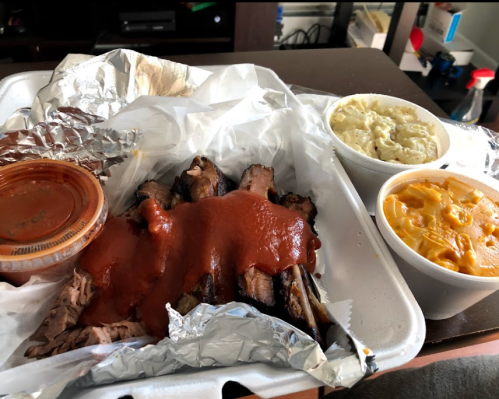A takeout container with sliced brisket topped with sauce, macaroni and cheese, and mashed potatoes on the side.