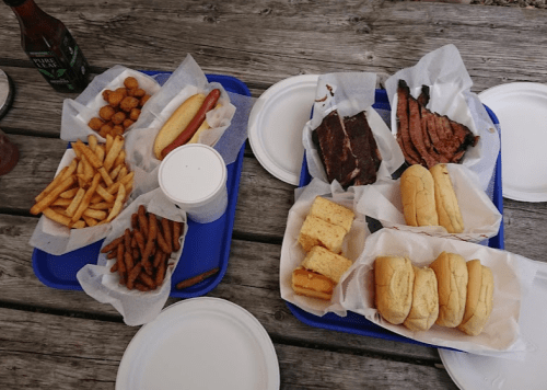 Two blue trays filled with various foods: fries, hot dog, brisket, cornbread, and buns, on a wooden table.