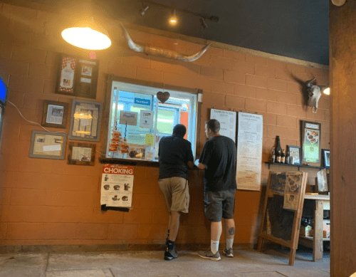 Two people stand at a service window in a rustic restaurant, with decor including a longhorn skull and various posters.