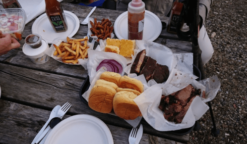 A picnic table spread with BBQ meats, buns, fries, onion slices, and sauces, alongside plates and utensils.