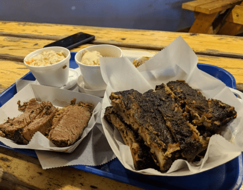 A blue tray with smoked meat, including brisket and ribs, alongside two cups of coleslaw on a wooden table.