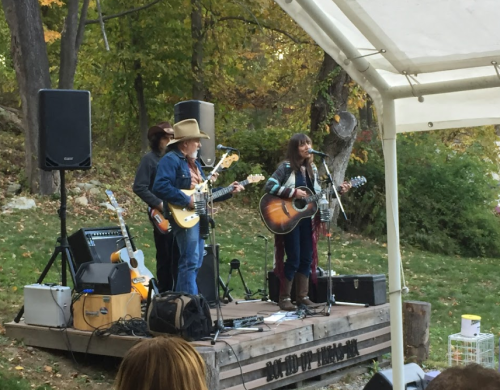 A band performs on an outdoor stage surrounded by trees, with a singer and musicians playing guitar and bass.