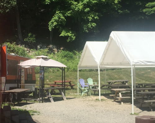 Outdoor seating area with picnic tables and tents, alongside a food truck in a green, wooded setting.