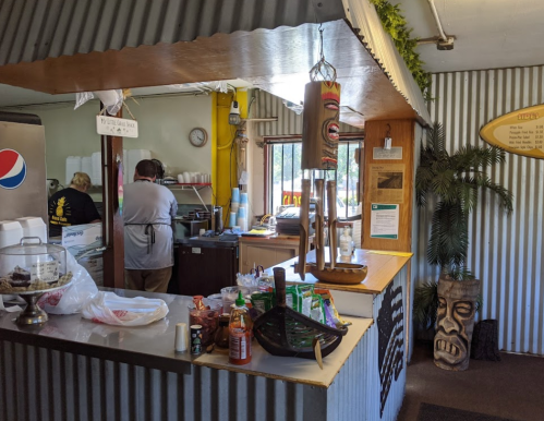 A cozy restaurant interior with a counter, staff preparing food, and tropical decor including a tiki statue.
