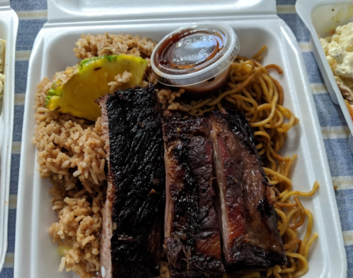 A takeout container with grilled ribs, rice, noodles, and a slice of lime, accompanied by a small cup of sauce.