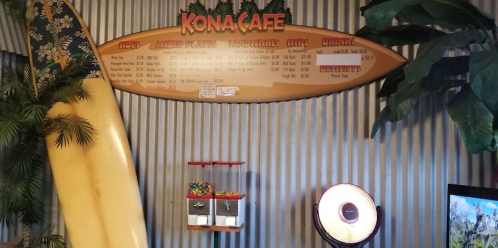 A menu board at Kona Cafe featuring food and drink options, with a large surfboard and tropical decor in the background.