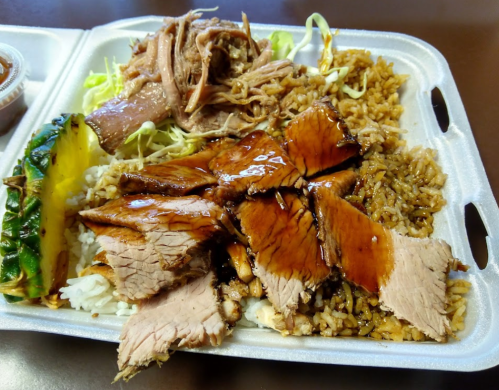 A takeout container filled with rice, pulled pork, sliced meat, and a piece of pineapple on a bed of lettuce.