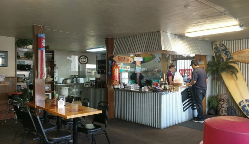 A cozy diner interior with a counter, surfboards, and patrons ordering food. Bright and casual atmosphere.