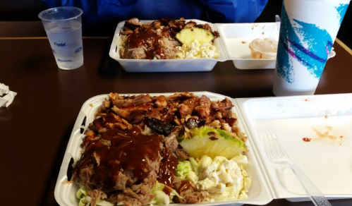 Two plates of barbecue food with pulled pork, chicken, pineapple, and sides, served with a drink and a small container of sauce.