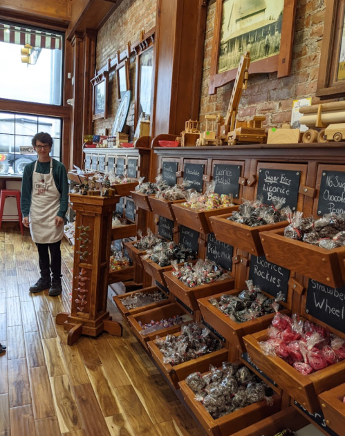 A candy shop interior with wooden shelves filled with various candies and a person in an apron standing nearby.