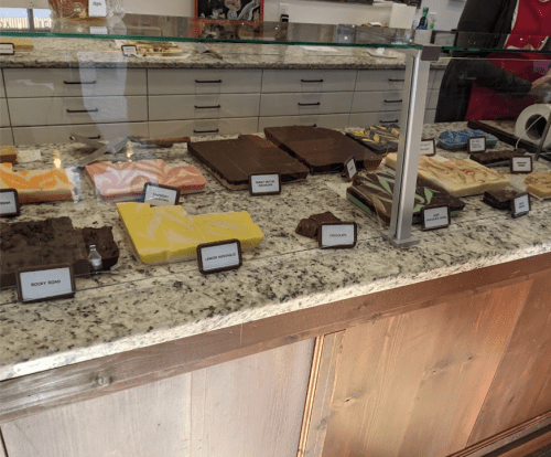 A display case filled with various types of fudge, labeled with names, on a granite countertop.