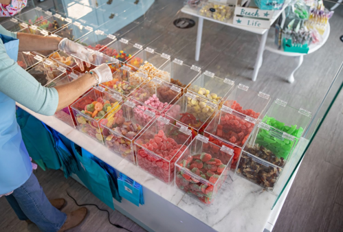 A person in an apron sorts colorful candies in clear containers at a candy shop.
