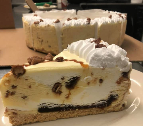 A slice of creamy cheesecake topped with whipped cream and chocolate bits, with the whole cake in the background.