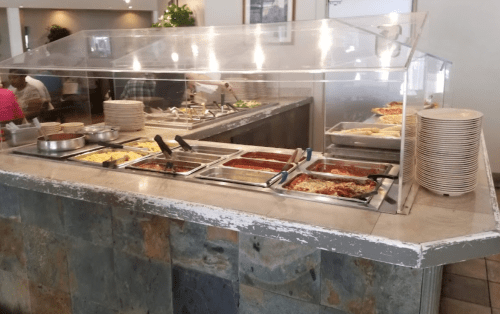 A buffet station with various food trays, including pasta and salads, under a glass cover in a dining area.