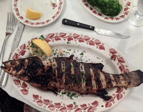 Grilled whole fish on a decorative plate, garnished with lemon and herbs, alongside a knife and fork.