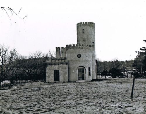 A small, stone tower structure with a round turret, surrounded by bare trees and a sandy ground, in black and white.
