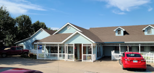 A light blue building with a brown roof, featuring white railings and a red car parked in front. Clear blue sky above.