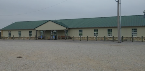 A single-story building with a green metal roof, surrounded by gravel and wooden fencing, under a cloudy sky.