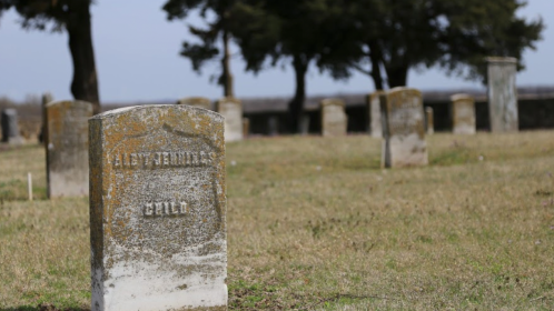 A weathered gravestone in a cemetery, surrounded by other gravestones and trees in the background.