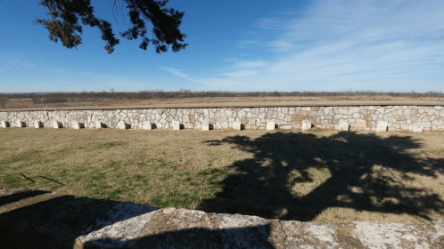 A stone wall stretches across a grassy field under a clear blue sky, with distant trees on the horizon.