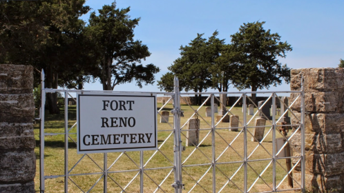 Gate to Fort Reno Cemetery, with gravestones visible in the background and trees lining the area under a clear blue sky.