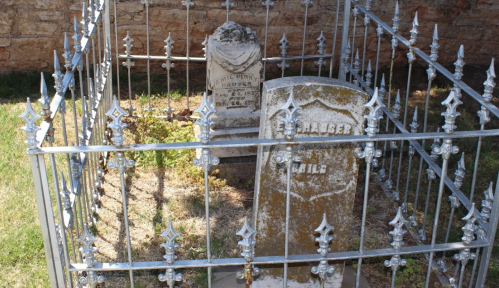 Two old gravestones enclosed in a decorative metal fence, surrounded by grass.