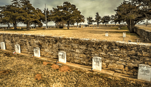 A cemetery with white gravestones lined along a stone wall, surrounded by trees and a cloudy sky.