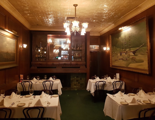 Elegant restaurant interior with wooden walls, chandeliers, and tables set with white linens and wine glasses.