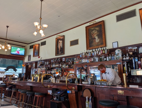 A bar with a wooden counter, shelves of liquor, a bartender, and a TV showing a sports game in a cozy setting.