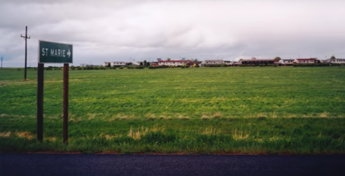 A green field with a signpost for "St Marie" and a distant view of houses under a cloudy sky.