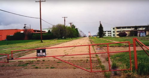 A red gate blocks a deserted road lined with power poles, leading to abandoned buildings under a cloudy sky.