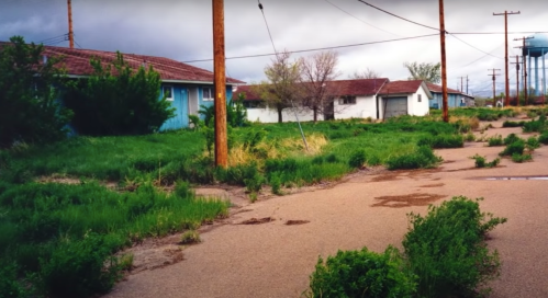 Abandoned buildings with overgrown grass and a cloudy sky, along a deserted road lined with power lines.