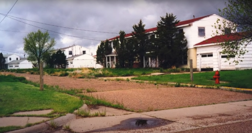 Abandoned buildings line a gravel road, with overgrown grass and a fire hydrant visible under a cloudy sky.