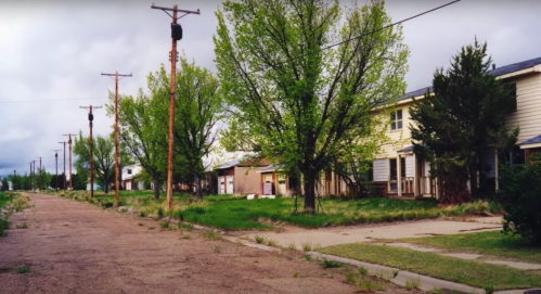 Abandoned street lined with overgrown grass, trees, and old houses under a cloudy sky.