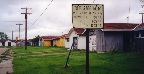 A weathered bus stop sign in a deserted area, with rundown houses and overgrown grass in the background.