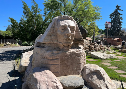A large stone sculpture of a sphinx with a human face, surrounded by rocks and greenery in a sunny outdoor setting.