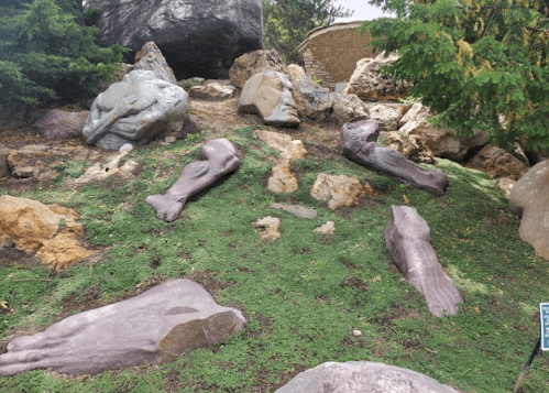 Sculptures of human limbs and heads scattered on grassy terrain, surrounded by rocks and trees.