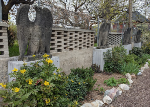 Four eagle statues on concrete pedestals, surrounded by greenery and flowers, against a textured wall.