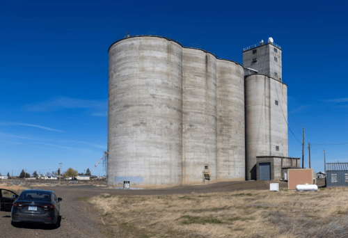 A large concrete grain silo stands against a clear blue sky, with a parked car nearby on a gravel path.
